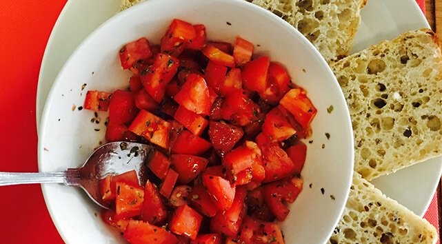 A bowl of cut tomatoes with olive oil and oregano placed next to three pieces of toasted baguette.