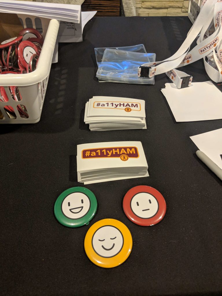 Stickers with #a11yHAM written on them, along with three buttons: green with an open smiley face, a yellow one with a smiley face, and a red one with a stern face.