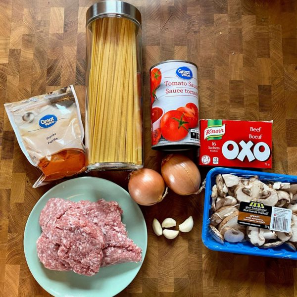 Top view of ground pork on a plate, a bag of paprika, a jar with spaghetti, a can of tomato sauce, two onions, some garlic cloves, a box of beef bouillon, and a package of mushrooms.