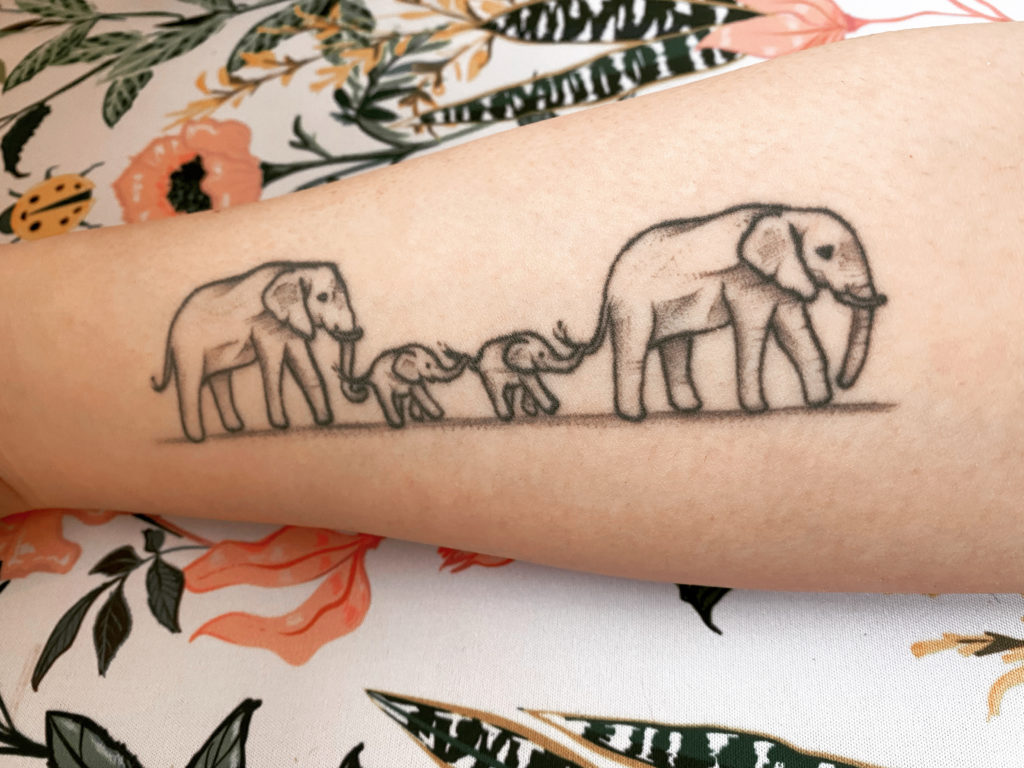 My right forearm is laying on a floral mat. My forearm has a large tattoo that consists of four elephants standing in a row. The elephants on the ends are adult elephants, with the two in the middle being baby elephants.