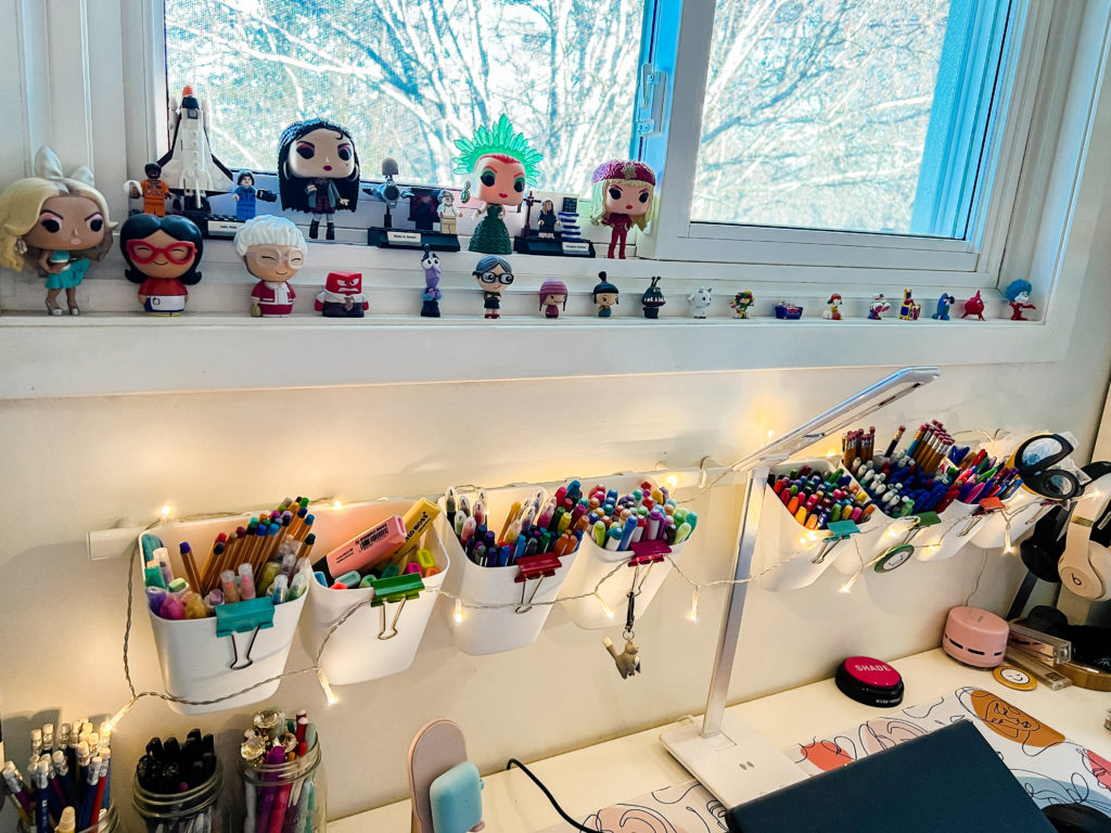 My figurines are on a window sill, and below is a railing with white containers holding various pens, pencils, highlighters and markers.