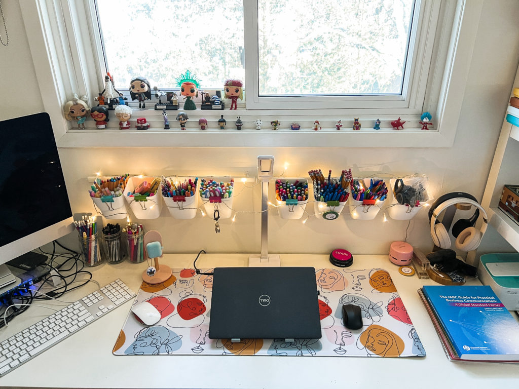 My desk with a large colourful mousepad, an iMac, a laptop and some books on it. Additionally, on the window sill above the desk rest a bunch of little figurines.