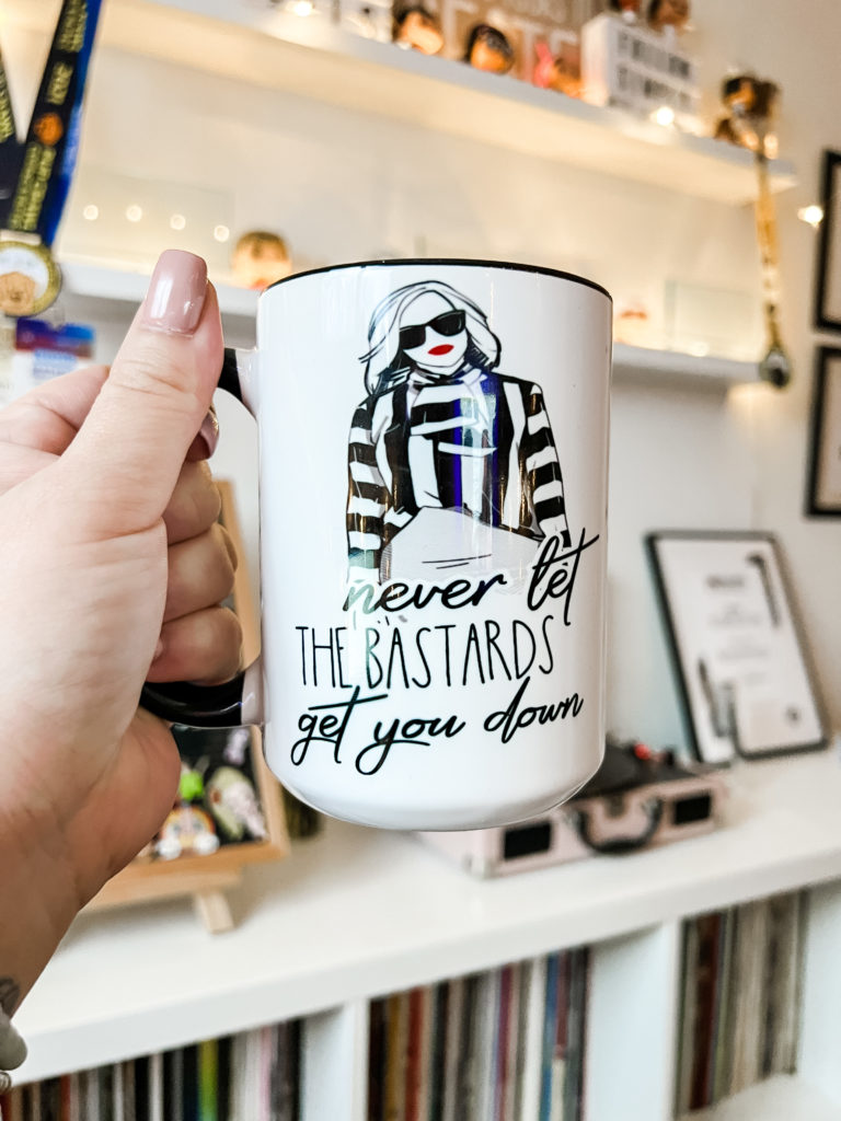 I'm holding up a mug with an illustration of Moira Rose and the words: Never let the bastards get you down.
