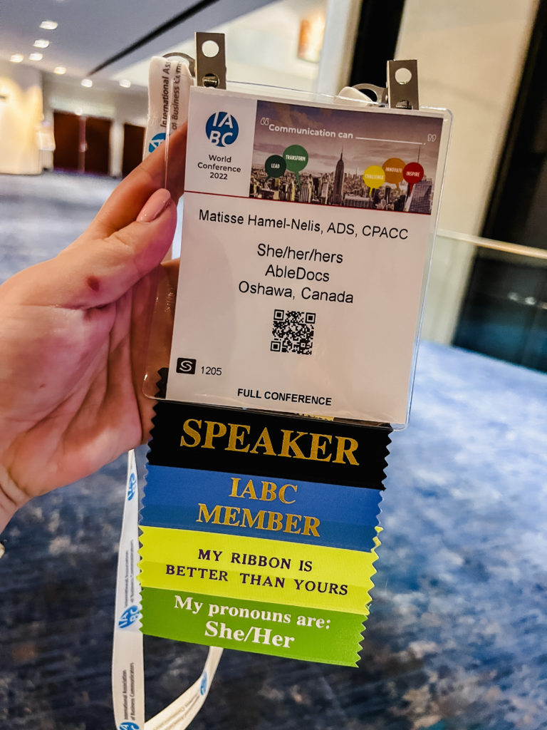 Matisse’s hand holds her IABC World Conference badge with her name, pronouns, company and location. Attached to the badge are four colourful ribbons. From top to bottom, they read: Speaker, IABC member, My ribbon is better than yours, and My pronouns are She/Her.