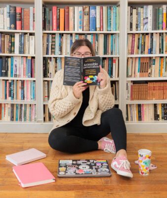 Matisse holds a copy of her book up in front of her face while sitting on a wooden floor in front of a bookcase. Her laptop, notebooks and a colourful mug surround her. 